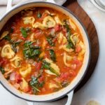 A white Dutch oven filled with hearty white bean tortellini soup with spinach and diced tomatoes.