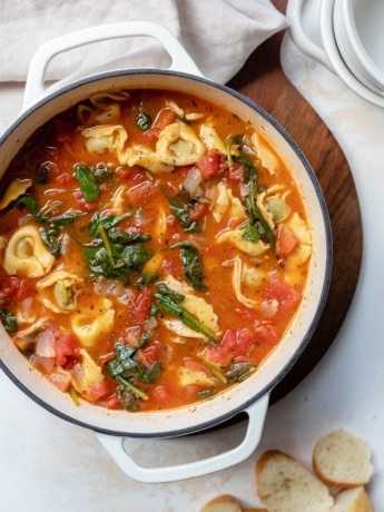 A white Dutch oven filled with hearty white bean tortellini soup with spinach and diced tomatoes.