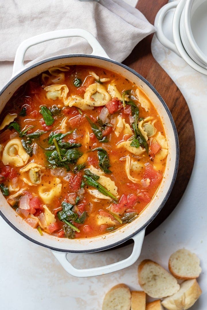 A white Dutch oven filled with hearty white bean tortellini soup with spinach and diced tomatoes.