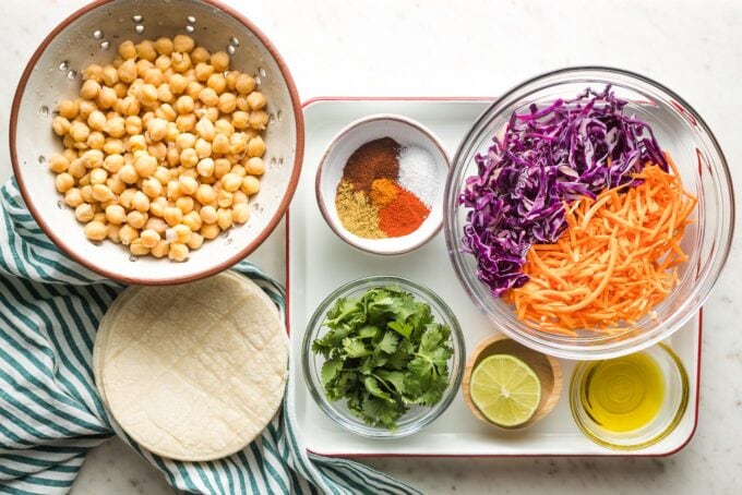Prep bowls filled with rinsed chickpeas, red cabbage, shredded carrots, seasonings, cilantro, lime, olive oil, and tortillas.