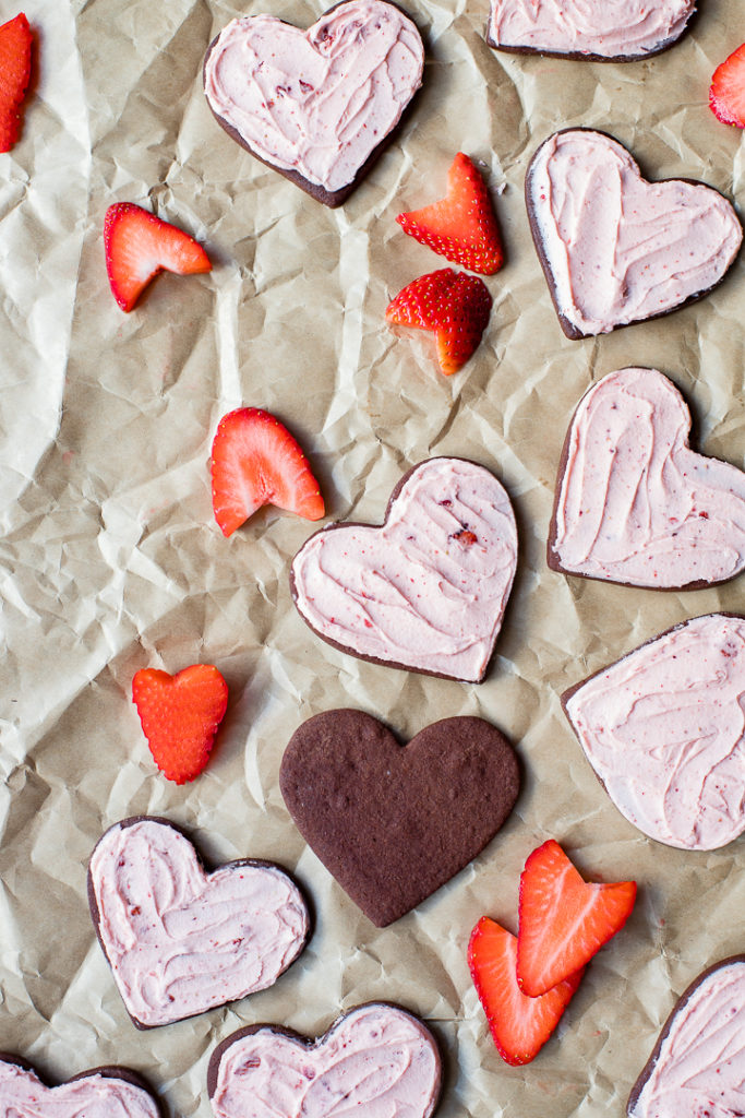 Heart-shaped strawberry-frosted chocolate sugar cookies, scattered with freshly-cut strawberries on crinkled parchment paper.