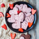 Heart-shaped strawberry-frosted chocolate sugar cookies on a black plate, scattered with freshly-cut strawberries.