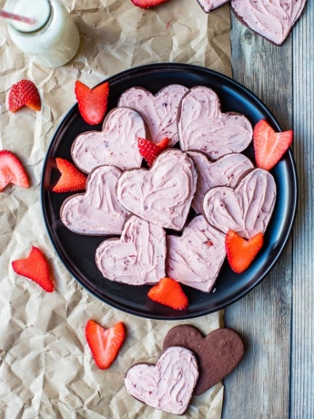 Heart-shaped strawberry-frosted chocolate sugar cookies on a black plate, scattered with freshly-cut strawberries.