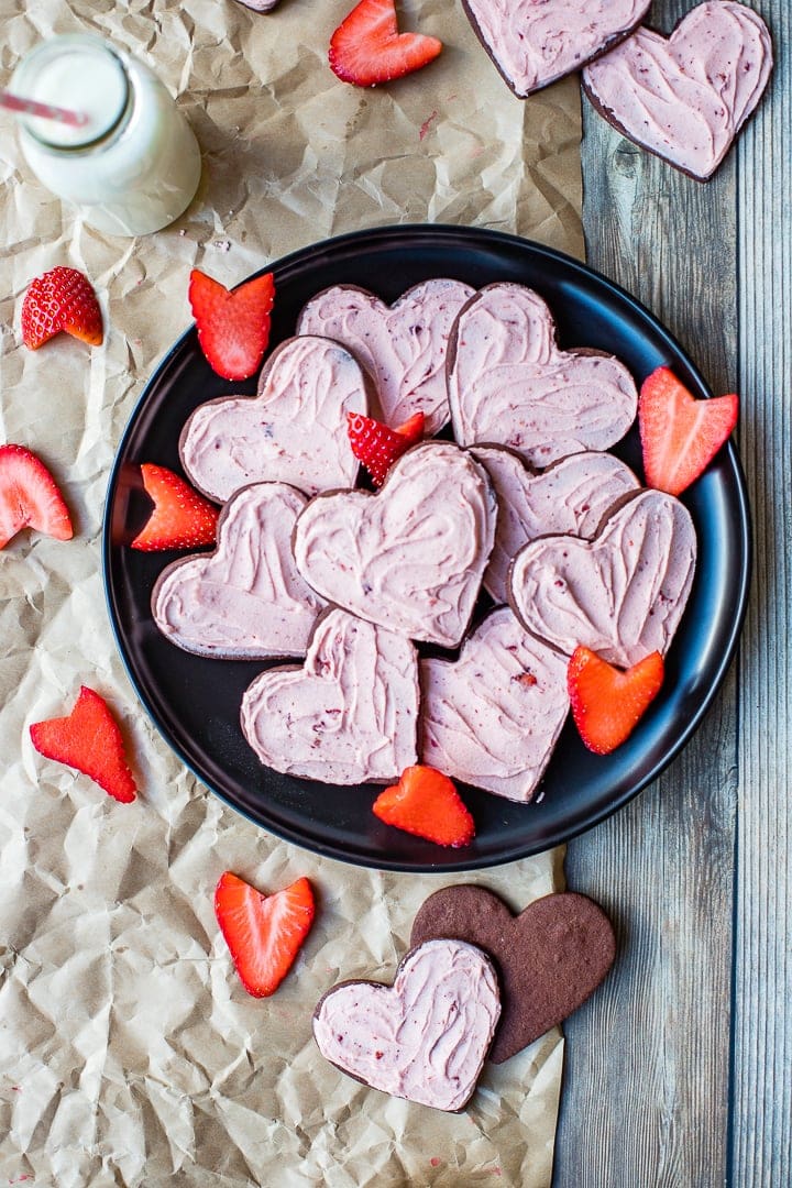 Heart-shaped strawberry-frosted chocolate sugar cookies on a black plate, scattered with freshly-cut strawberries.