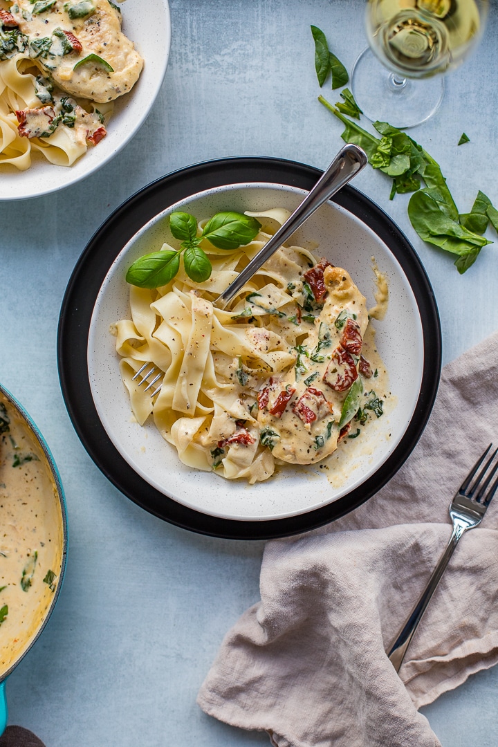 A table scene with plates of creamy Tuscan chicken served with pasta, fresh herbs, and white wine.