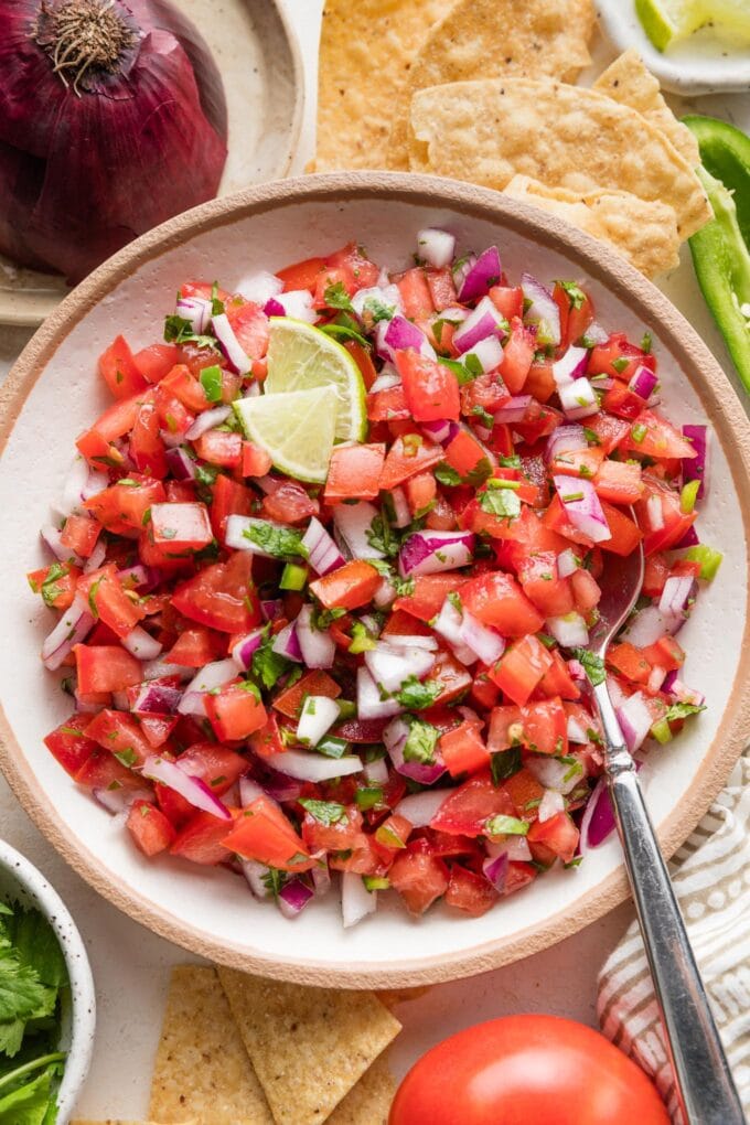 Bowl of homemade pico de gallo surrounded by tortilla chips, extra onion, and extra cilantro.