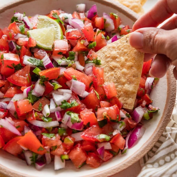 Woman's hand using a tortilla chip to scoop freshly homemade pico de gallo out of a bowl.