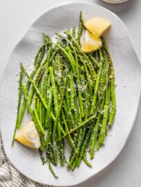 Oval ceramic plate piled with roasted lemon asparagus served with Parmesan and extra lemon wedges.