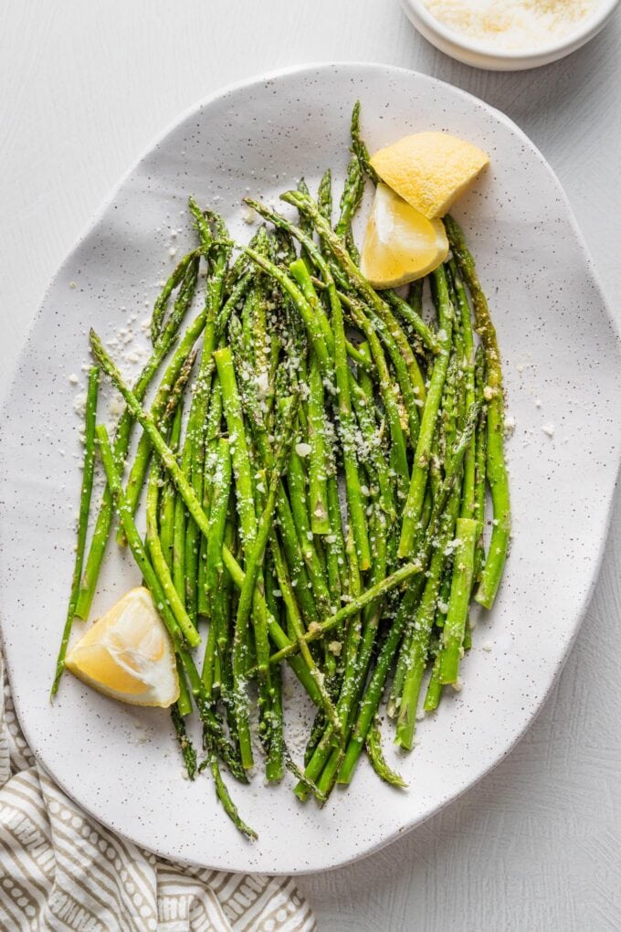 Oval ceramic plate piled with roasted lemon asparagus served with Parmesan and extra lemon wedges.