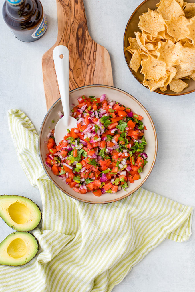 A bowl full of homemade pico de gallo, with chips, avocado, and a cold beer.