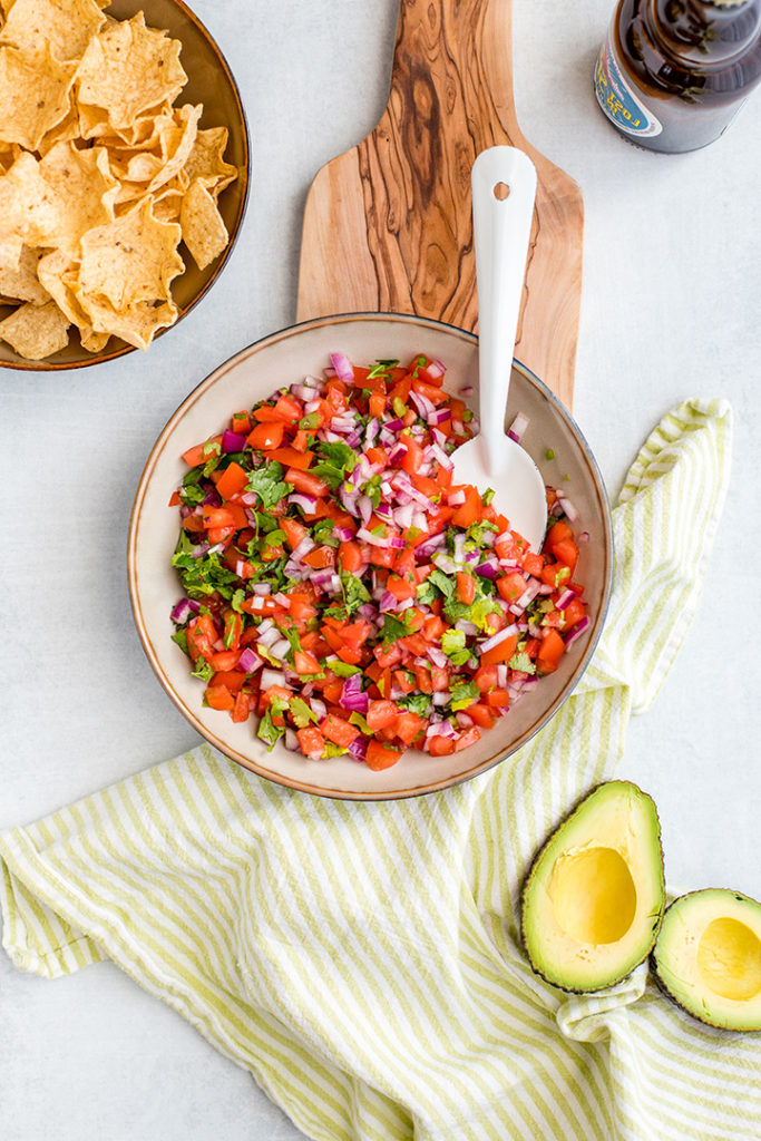 A bowl full of homemade pico de gallo, with chips, avocado, and a cold beer.