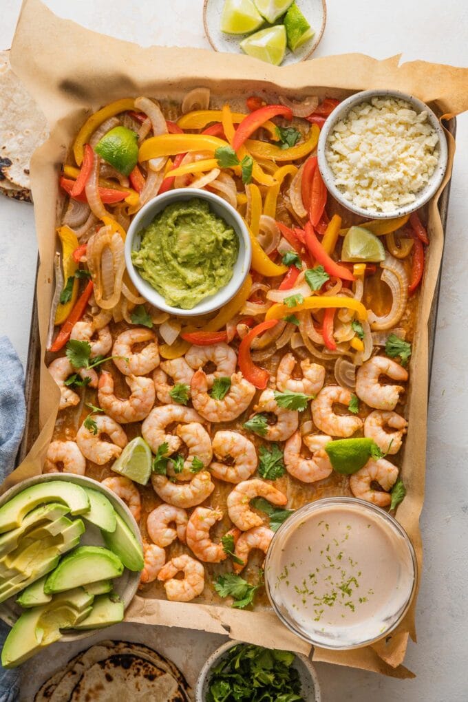 Large sheet pan filled with shrimp and fajita veggies, with small bowls scattered nearby with crumbled Cotija, a chipotle lime crema, sliced avocado, guacamole, tortillas, and lime wedges.