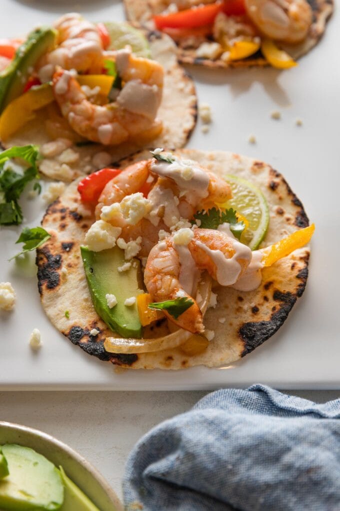 Angled close up of a shrimp fajita served in a warm, charred tortilla with sliced avocado and a chipotle crema drizzled on top.