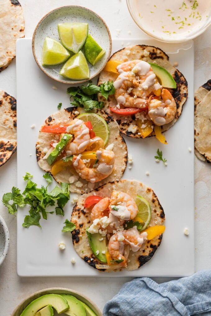 Platter with three shrimp fajitas with peppers, onions, crema, avocado, and fresh cilantro leaves.