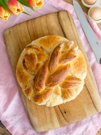 Flat-lay of a loaf of homemade paska with a cross on top, with tulips and eggs in the background.