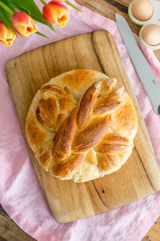 Flat-lay of a loaf of homemade paska with a cross on top, with tulips and eggs in the background.