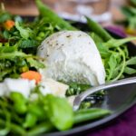 Close-up of a cut-open ball of fresh Buffalo mozzarella cheese nestled in a bright salad of arugula and sugar snap peas.