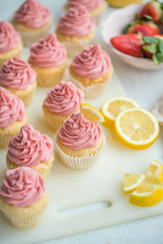 An array of strawberry lemonade cupcakes scattered next to fresh strawberries and cut lemon slices.