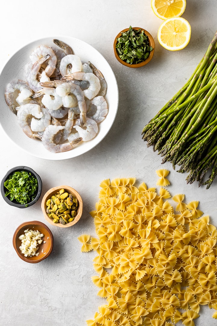Flat lay of raw shrimp, lemons, herbs, spices, asparagus, and farfalle (bowtie) pasta.