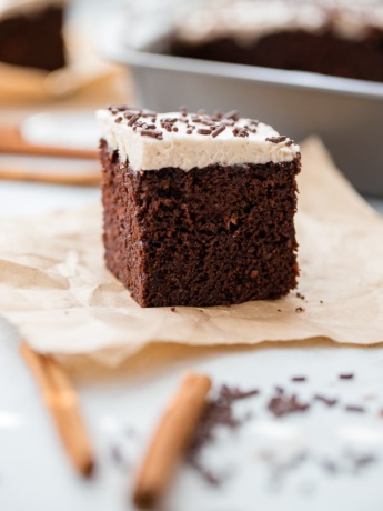 A small square slice of Mexican chocolate cake with cinnamon frosting and chocolate sprinkles, set on a piece of parchment paper.