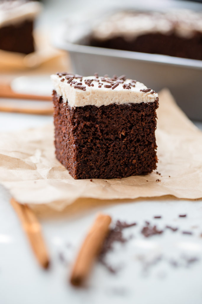 A small square slice of Mexican chocolate cake with cinnamon frosting and chocolate sprinkles, set on a piece of parchment paper.