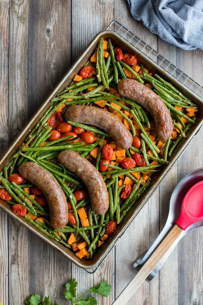 Flat-lay of an Italian sausage sheet pan dinner with sweet potatoes, green beans, and cherry tomatoes, resting on a cooling rack with a red serving spoon to the side.