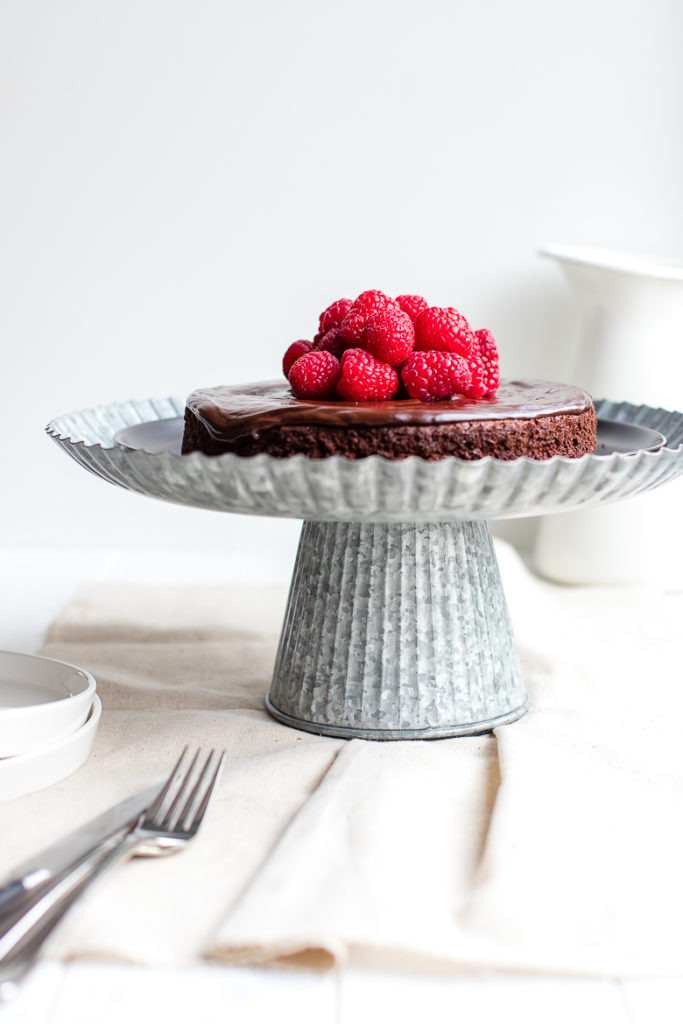 A perfect flourless chocolate cake presented on a pretty galvanized cake stand, topped with fresh raspberries.