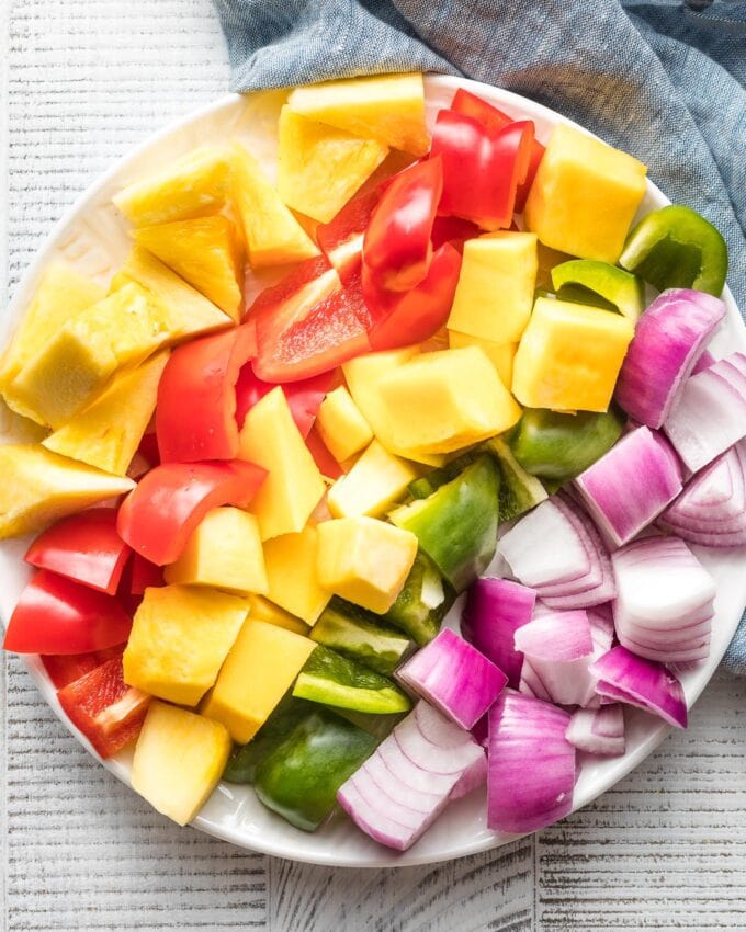 Cubed mango, red bell pepper, pineapple, green bell pepper, and red onion lined up on a plate, ready to be threaded onto kabobs.