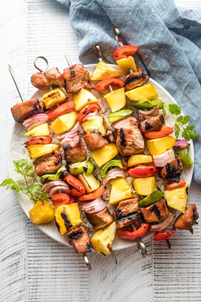Kabobs with pork, pineapple, mango, onions, and peppers piled on a white plate.