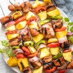 Kabobs with pork, pineapple, mango, onions, and peppers piled on a white plate.