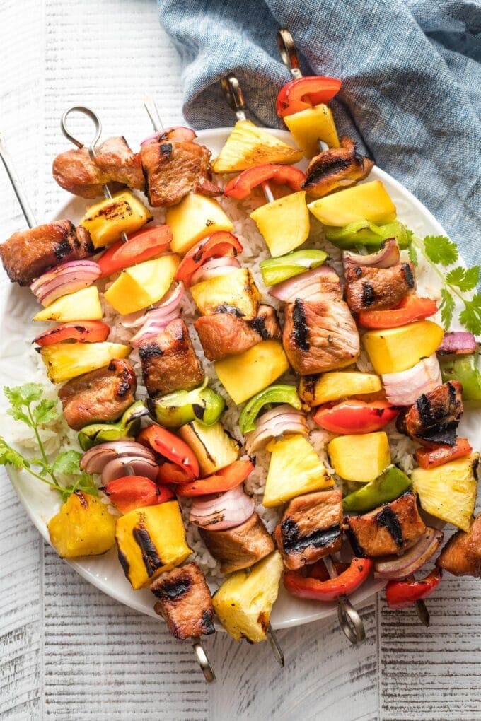 Kabobs with pork, pineapple, mango, onions, and peppers piled on a white plate.