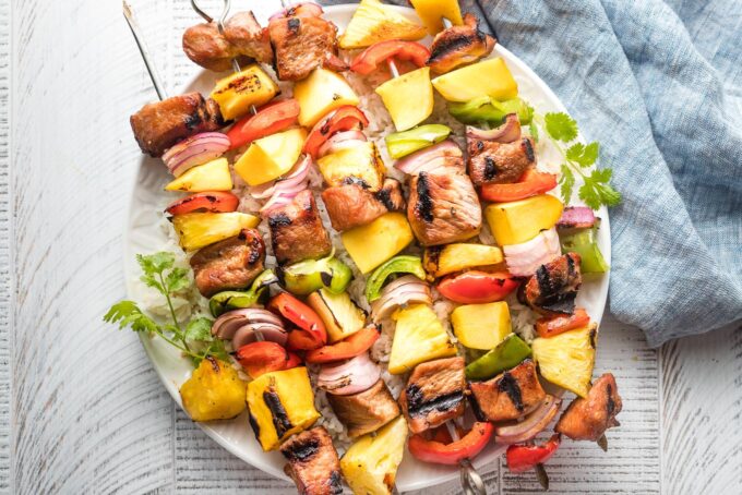 Kabobs with pork, pineapple, mango, onions, and peppers piled on a white plate.