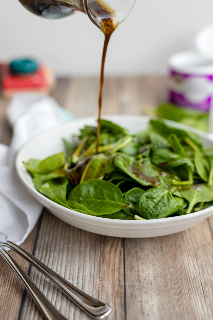 Pouring balsamic vinaigrette out of a cruet onto a simple spinach salad.