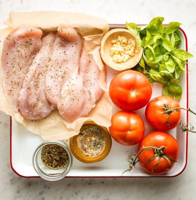 Tray with chicken breasts, minced garlic, fresh basil, tomatoes, olive oil, balsamic vinegar, and dried seasonings.