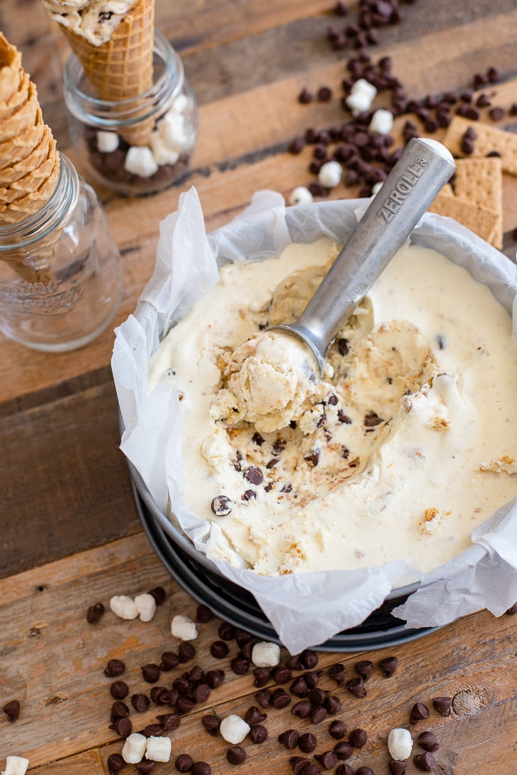 No-churn s'mores ice cream in a large round container with chocolate, marshmallows, and ice cream cones scattered around.