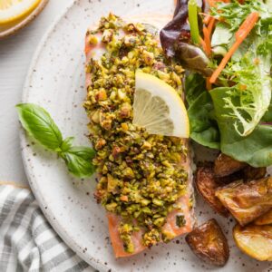 Plate with a large filet of salmon coated with a pistachio herb topping and a wedge of lemon, served with salad and roasted potatoes.