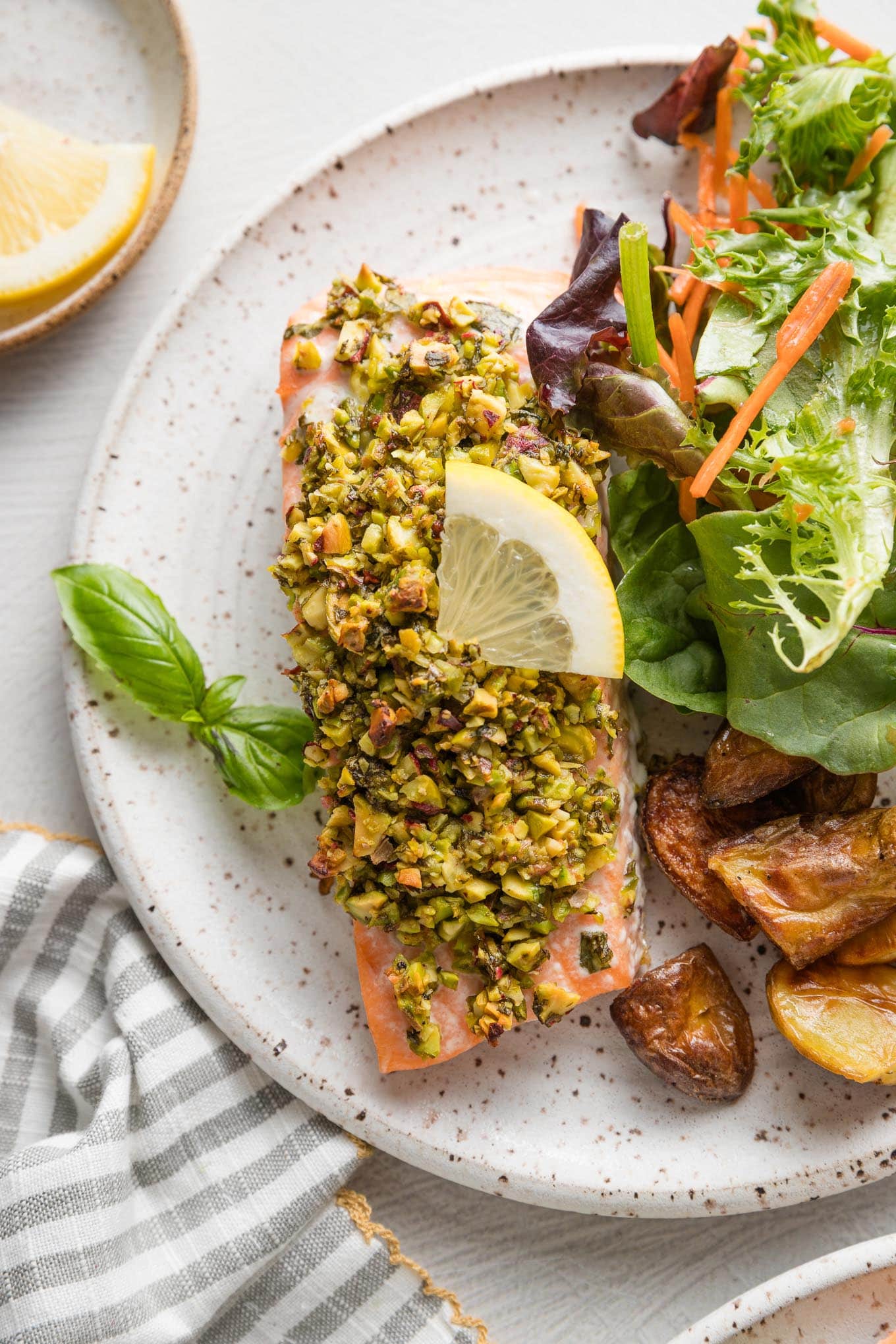 Plate with a large filet of salmon coated with a pistachio herb topping and a wedge of lemon, served with salad and roasted potatoes.