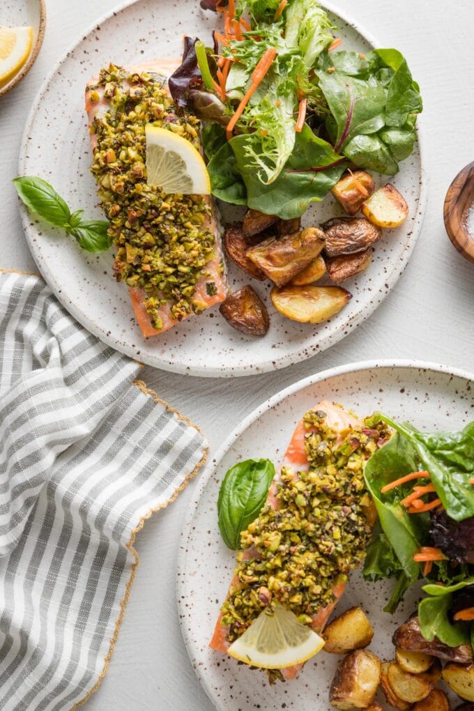 Two dinner plates with pan-seared pistachio herb salmon, roasted baby potatoes, and salad greens topped with Italian dressing.