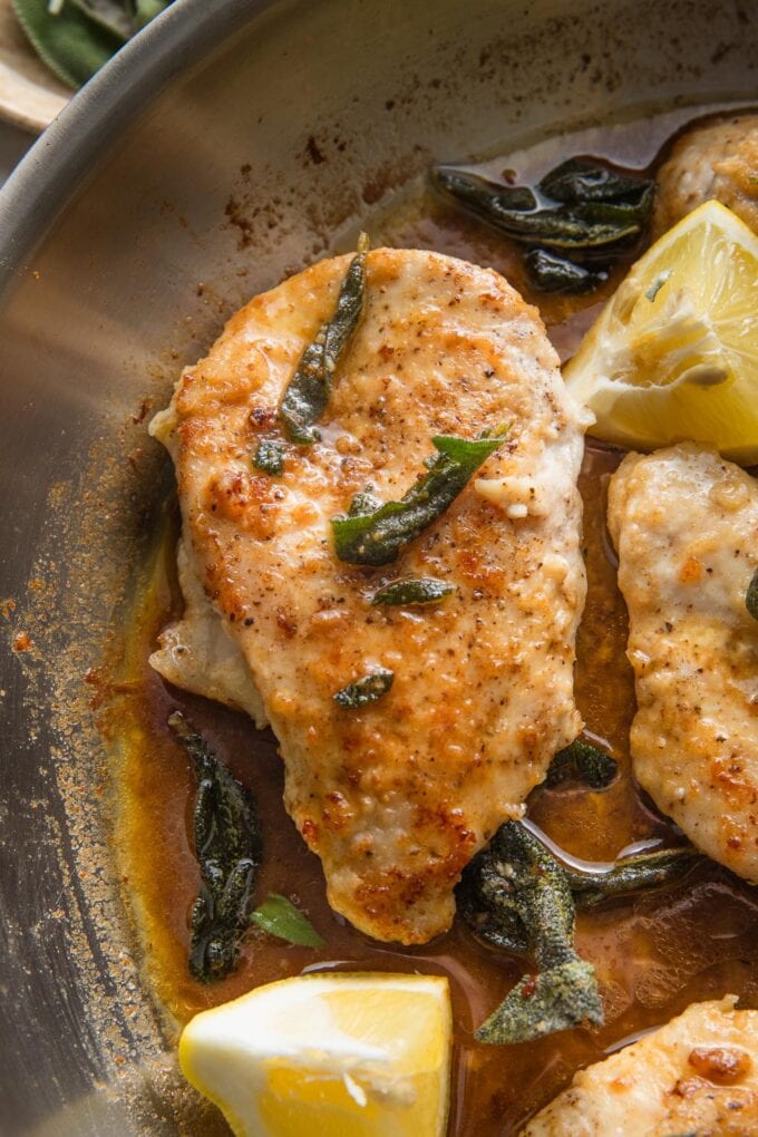 Close up of a brown butter sage chicken breast in a skillet with lemon wedges nearby.