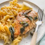 Small plate with a chicken breast cooked in a brown butter sage white sauce served with egg noodles and extra sage leaves.