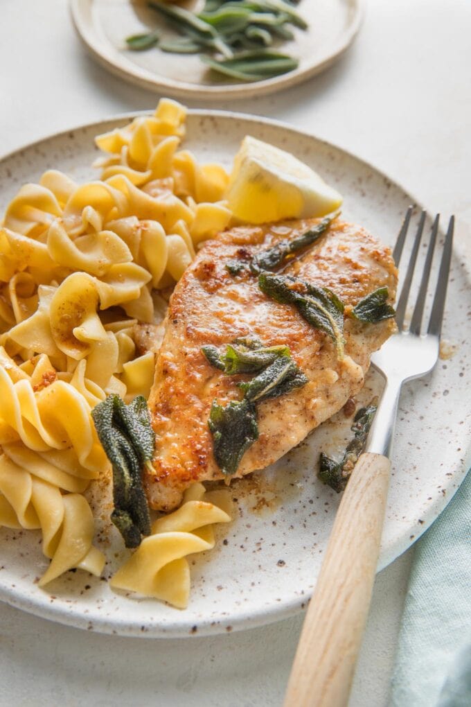 Small plate with a chicken breast cooked in a brown butter sage white sauce served with egg noodles and extra sage leaves.