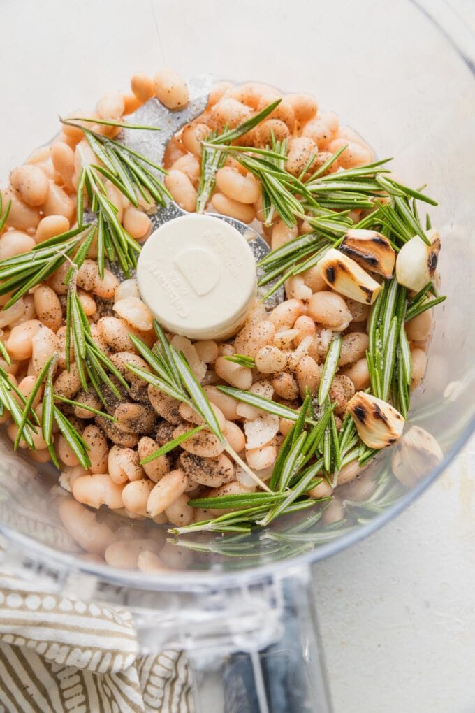 Bowl of a food processor holding cannellini beans, toasted whole garlic cloves, fresh rosemary sprigs, lemon juice, salt, and pepper.