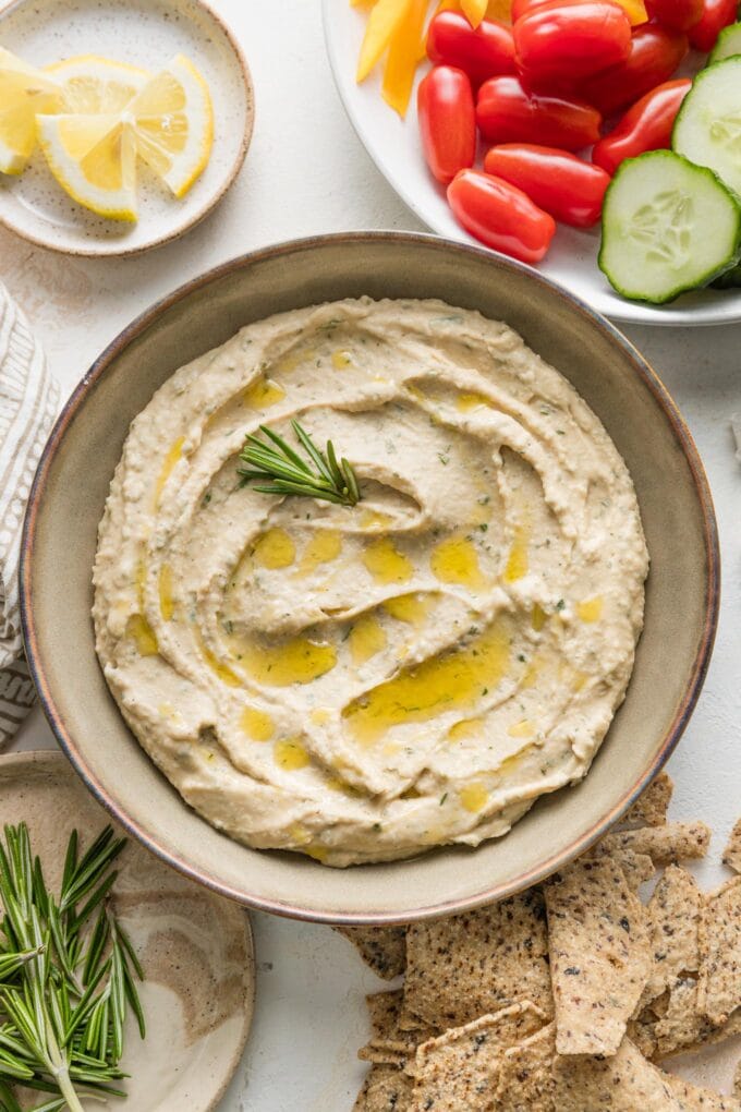 Overhead view of a bowl of Cannellini bean dip with garlic and rosemary, surrounded by tortilla chips and fresh veggies for dipping.