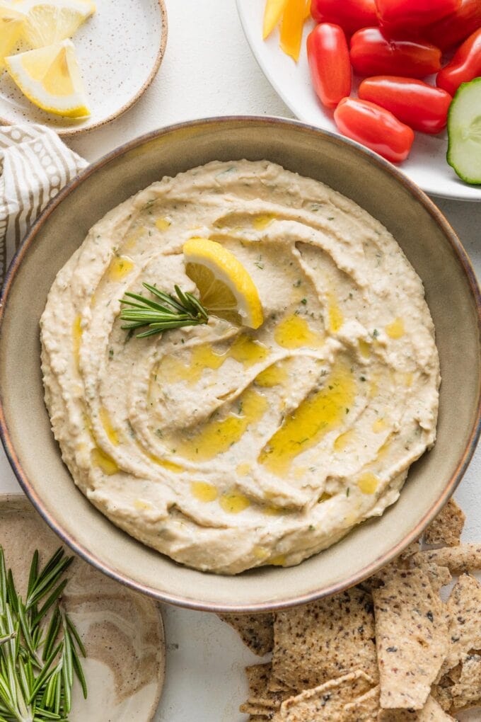 Bowl of rosemary white bean dip with toasted garlic, garnished with fresh rosemary, lemon wedges, and an extra drizzle of olive oil.