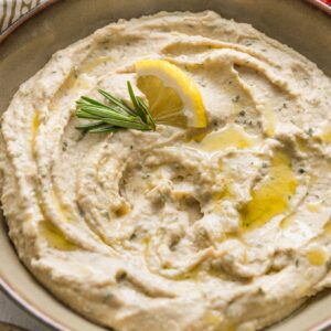Small bowl full of a creamy garlic rosemary white bean dip garnished with olive oil, fresh rosemary, and a lemon wedge.
