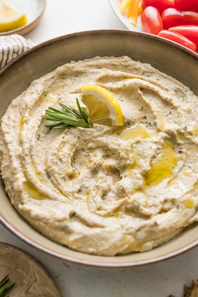 Small bowl full of a creamy garlic rosemary white bean dip garnished with olive oil, fresh rosemary, and a lemon wedge.