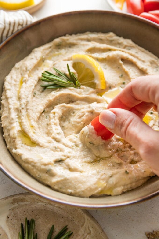 Woman's hand dipping a cherry tomato in a bowl of rosemary white bean dip.