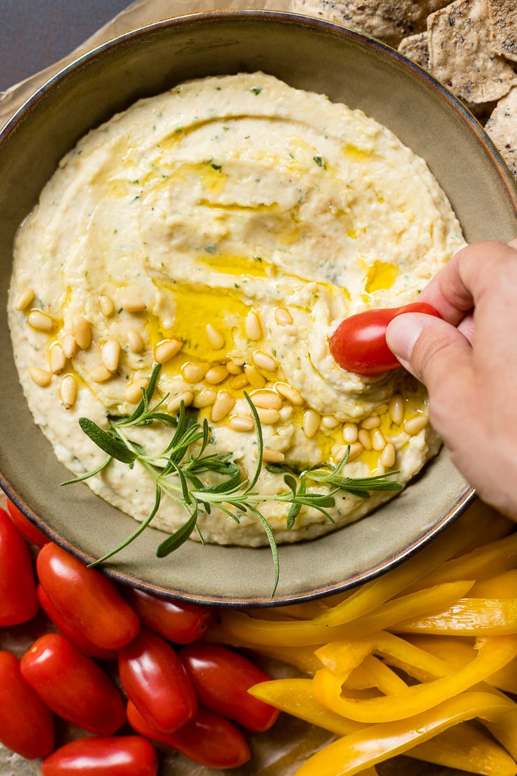 A hand dipping a cherry tomato into a bowl of garlic rosemary white bean dip.