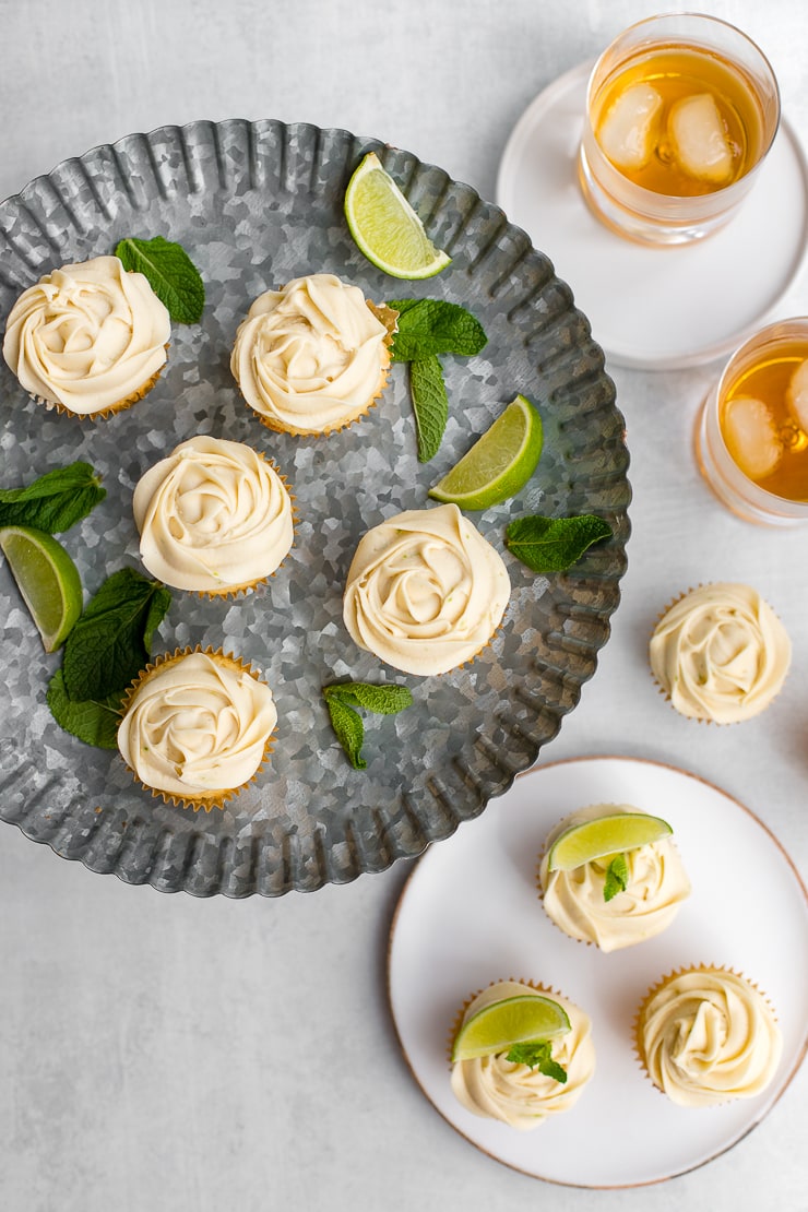 Flat-lay of mojito cupcakes arranged on a galvanized cake platter surrounded by more cupcakes and glasses of rum on the rocks.