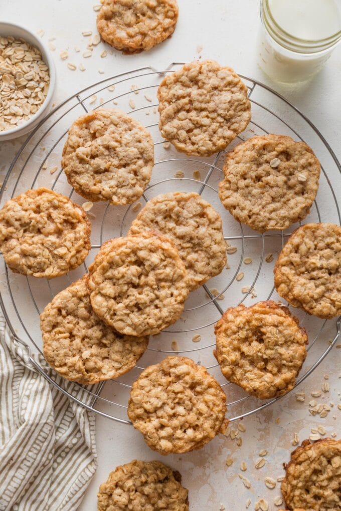 Old fashioned oatmeal cookies with chewy centers and lacy edges cooling on a small, round wire rack, with a small glass of milk and a bowl with extra old fashioned oats nearby.